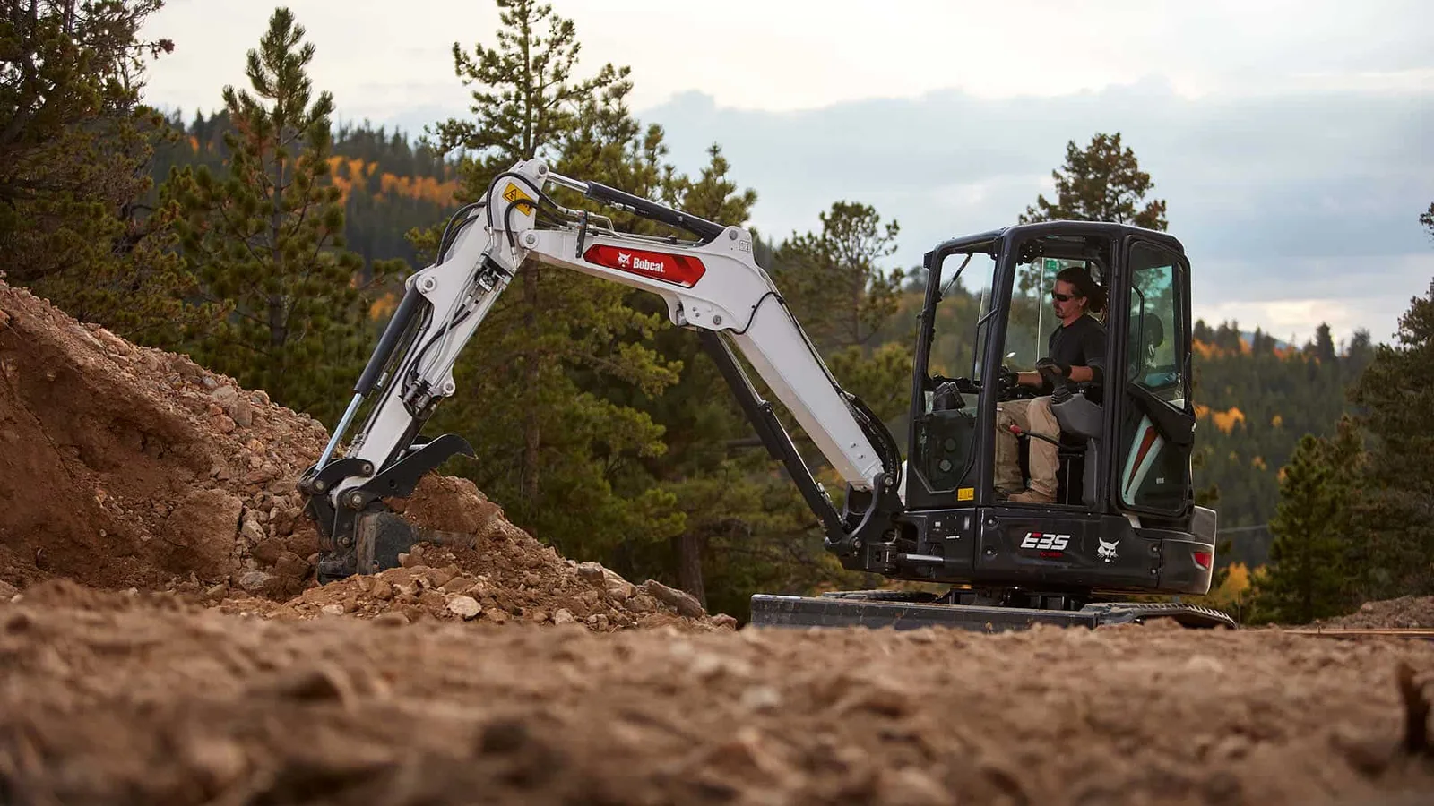 White Bobcat compact excavator digging into a rocky dirt bank with its bucket, with pine trees behind it