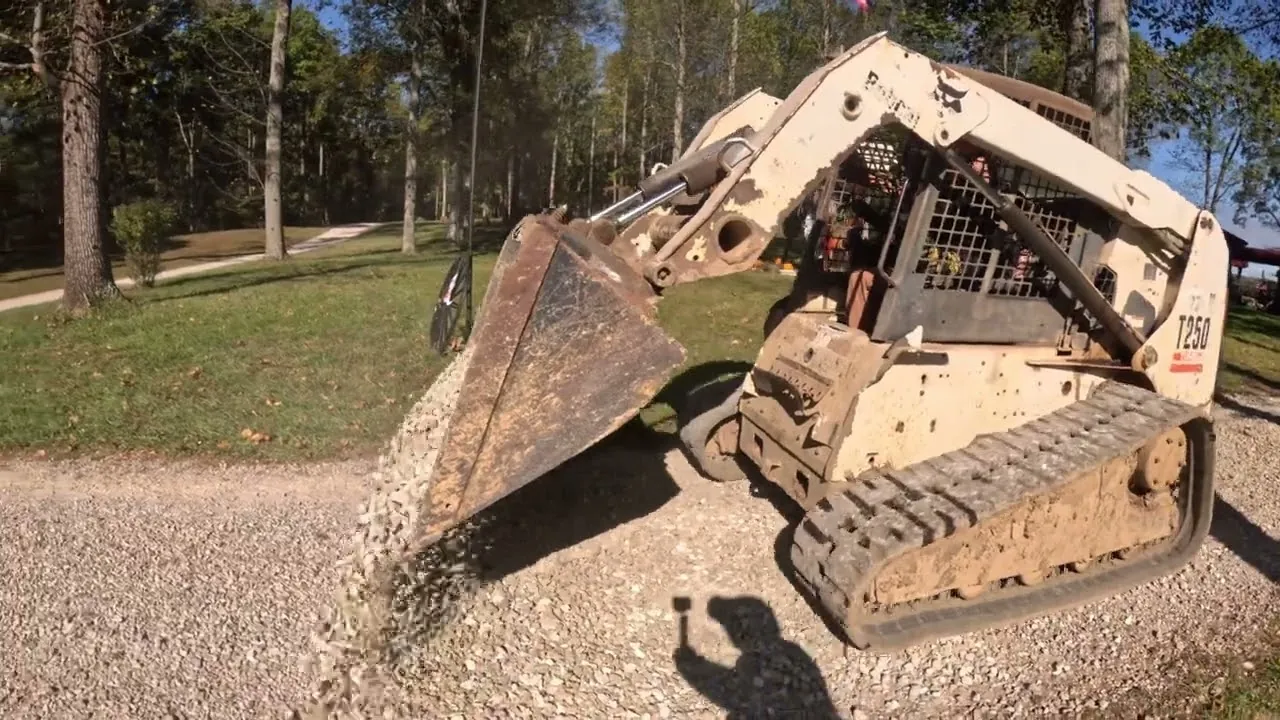 Bobcat compact track loader dumping a bucket of gravel onto a rural gravel drive, with trees and a lawn behind it