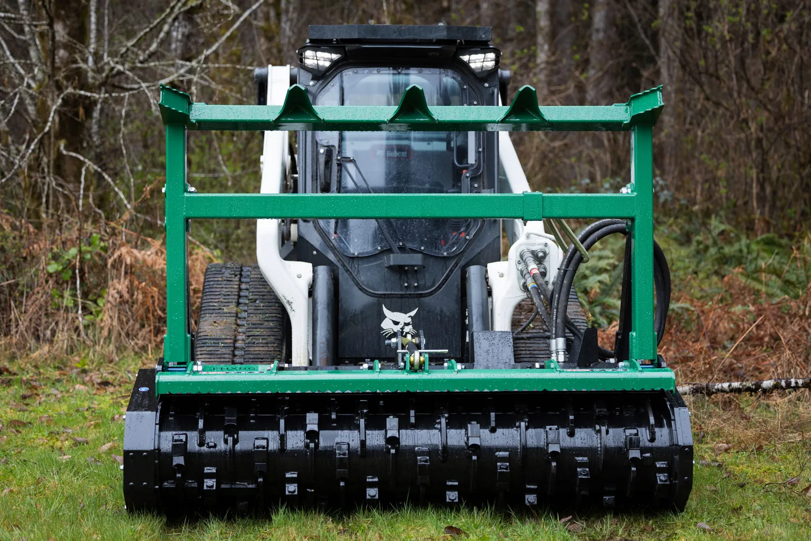 White Bobcat compact track loader with a green forestry mulcher head, parked on grass in front of brushy woods