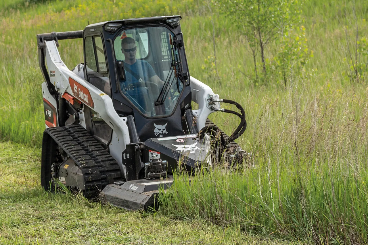 Tracked skid steer cutting tall grass and weeds in an overgrown field with a brush mower